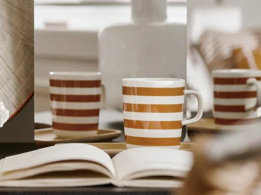 Der Oiva Tasaraita Becher mit Henkel von Marimekko in der Ambienteansicht. Das Wollweiß und das Braun gehen zusammen ein harmonisches Streifenmuster ein.