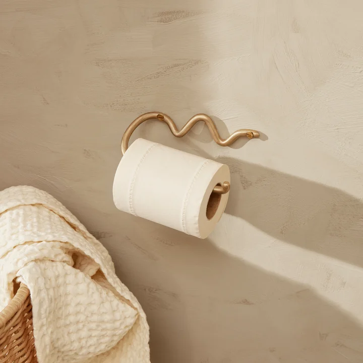 Der Curvature Toilettenpapierhalter von Ferm Living an einer hellen Steinwand neben einem Rattan-Korb