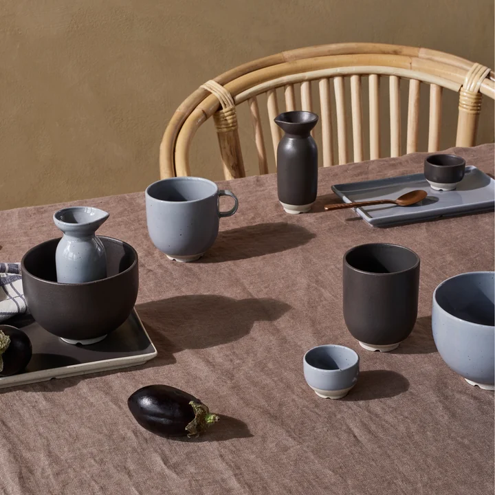 Das Eli Geschirr von Broste Copenhagen in soft blue und charoal auf dem Esstisch neben einem Rattan-Stuhl