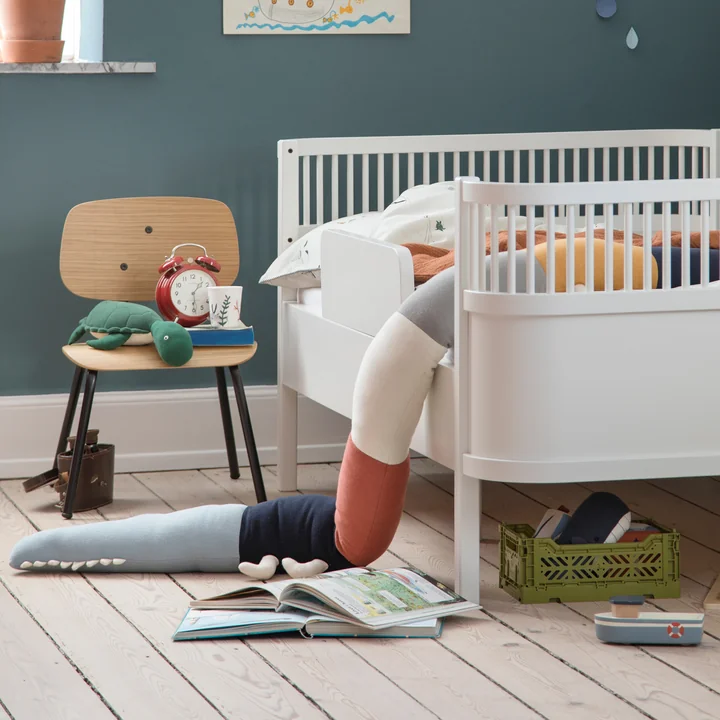 Das Sleepy Croc Kissen und der Oakee Stuhl von Sebra neben dem weißen Kinderbett